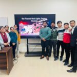 A cheerful group of MBA students and a faculty member standing in front of a presentation screen at Jaipuria Institute of Management, Ghaziabad, post an interactive learning session.