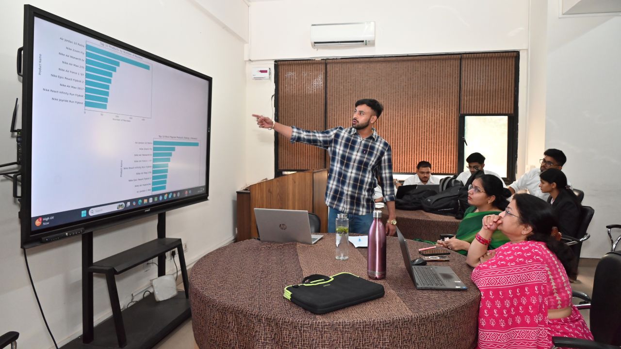 Man in a plaid shirt presenting data charts on a large screen to colleagues in a meeting room.