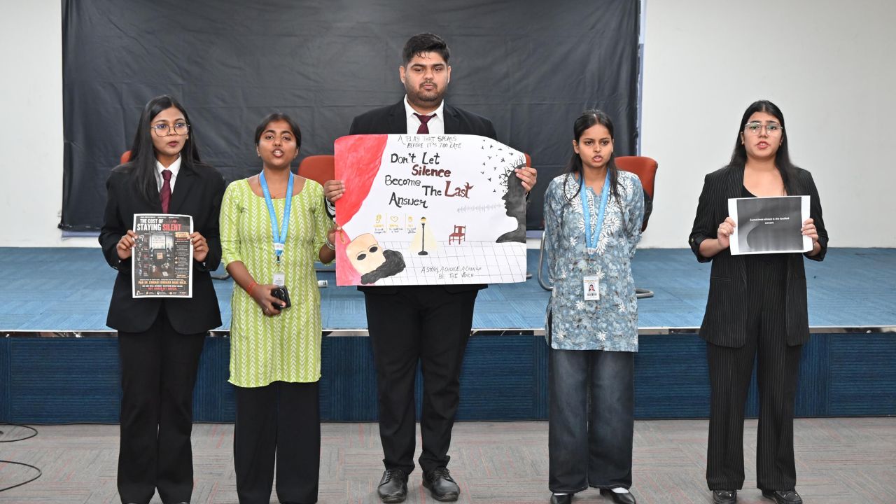 Five young adults stand on a stage, holding posters about speaking up and breaking silence. Center man in a suit holds a large poster with anti-silence message. The group is dressed in business casual!
