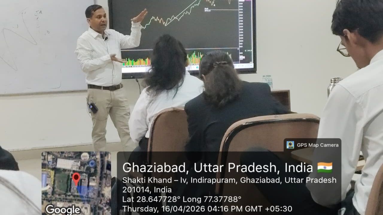 Instructor presenting stock-market charts to students in a classroom; monitor displays an uptrend graph with colorful bars beneath it.