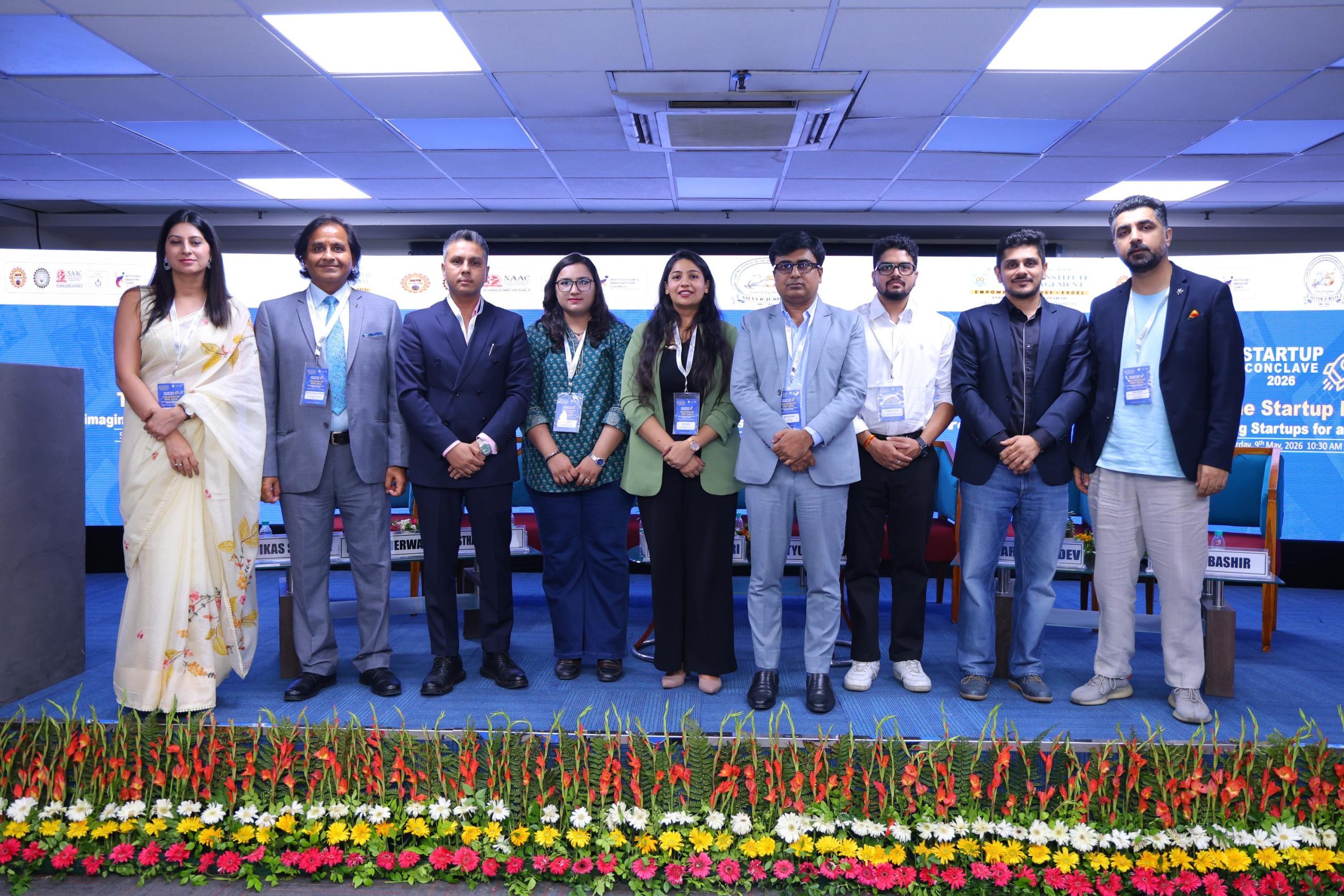Startup Conclave 2026 AI Driven Innovation 25 - Jaipuria Institute of Management, Indirapuram Ghaziabad Group of eight professionals standing on a stage at a conference, with colorful flower decorations in the foreground and banners in the background.