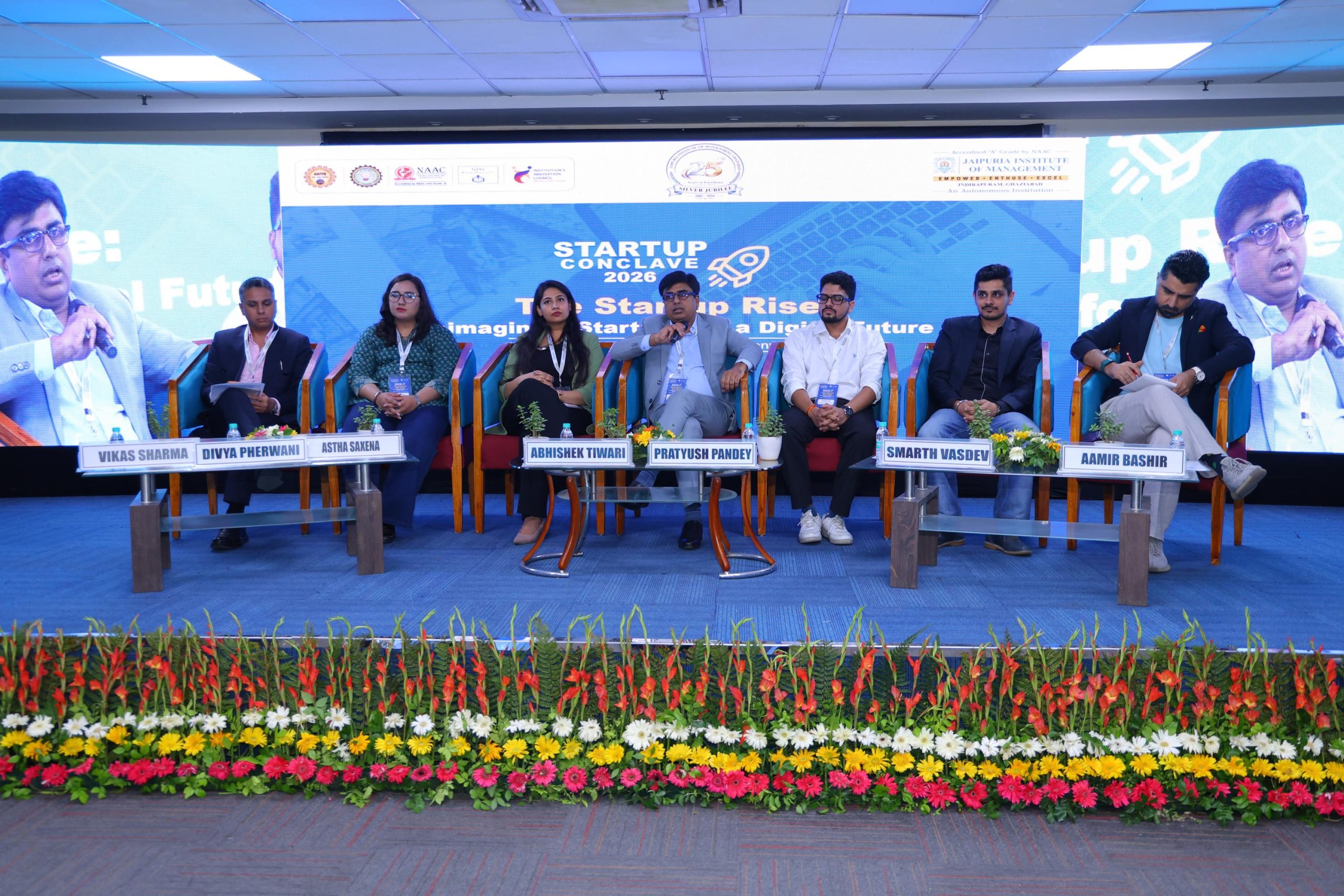 Startup Conclave 2026 AI Driven Innovation 9 - Jaipuria Institute of Management, Indirapuram Ghaziabad Panel of seven speakers seated on a blue stage with nameplates in front, startup conference backdrop reading 'Startup Conclave 2026' and decorative flowers in the foreground.