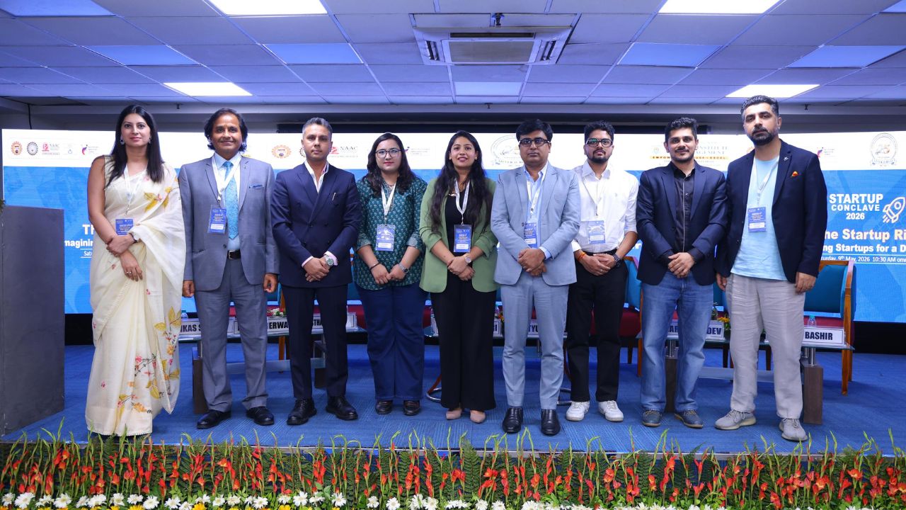 Group of ten professionals on a stage at a startup conclave, posing for a photo with badges visible under bright conference lights.