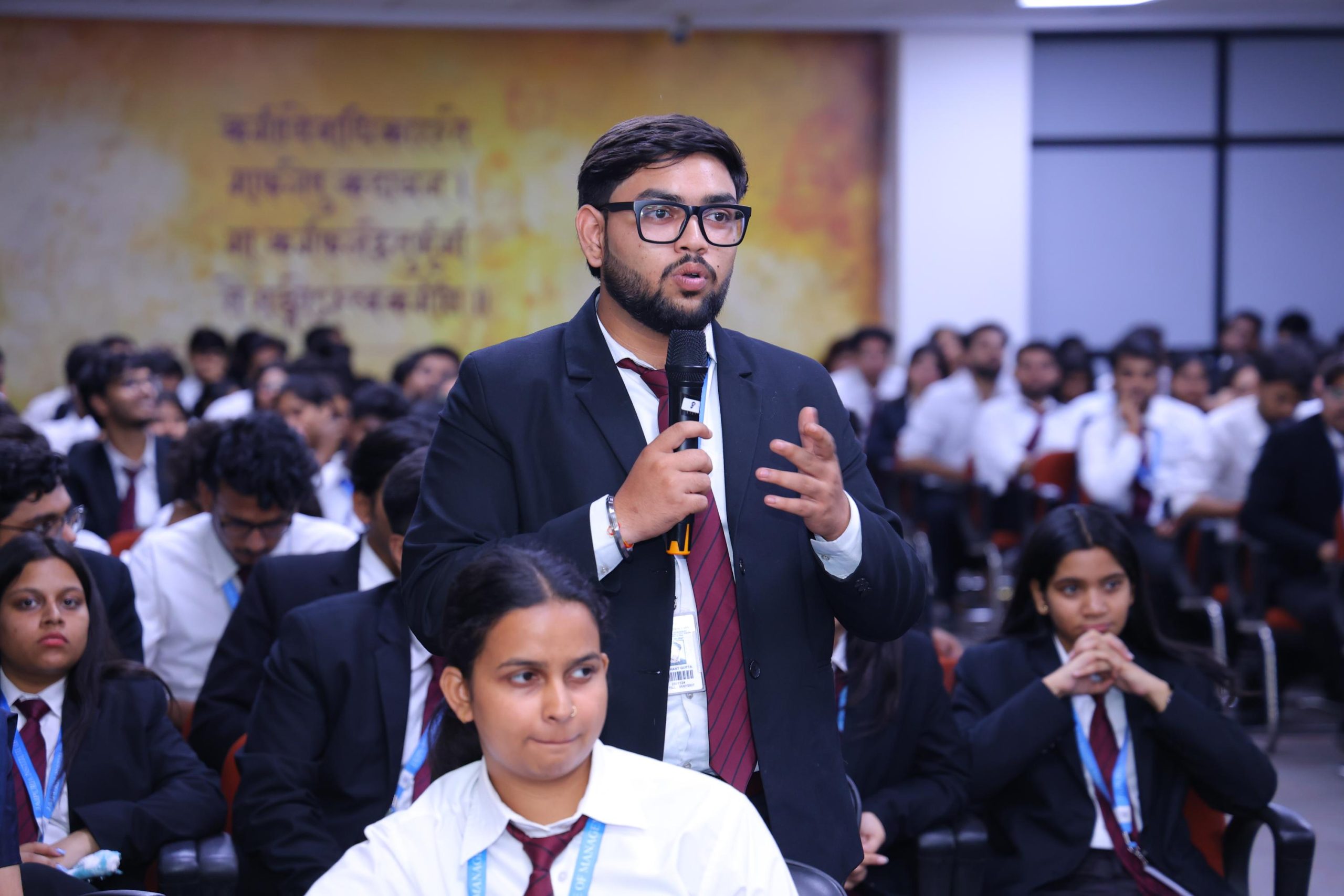 Startup Conclave 2026 Building Responsible Investment Ready Startups in the Digital Era 15 - Jaipuria Institute of Management, Indirapuram Ghaziabad Young man in a suit and glasses speaks into a handheld microphone at a student conference, with peers in white shirts and red ties seated in the background.