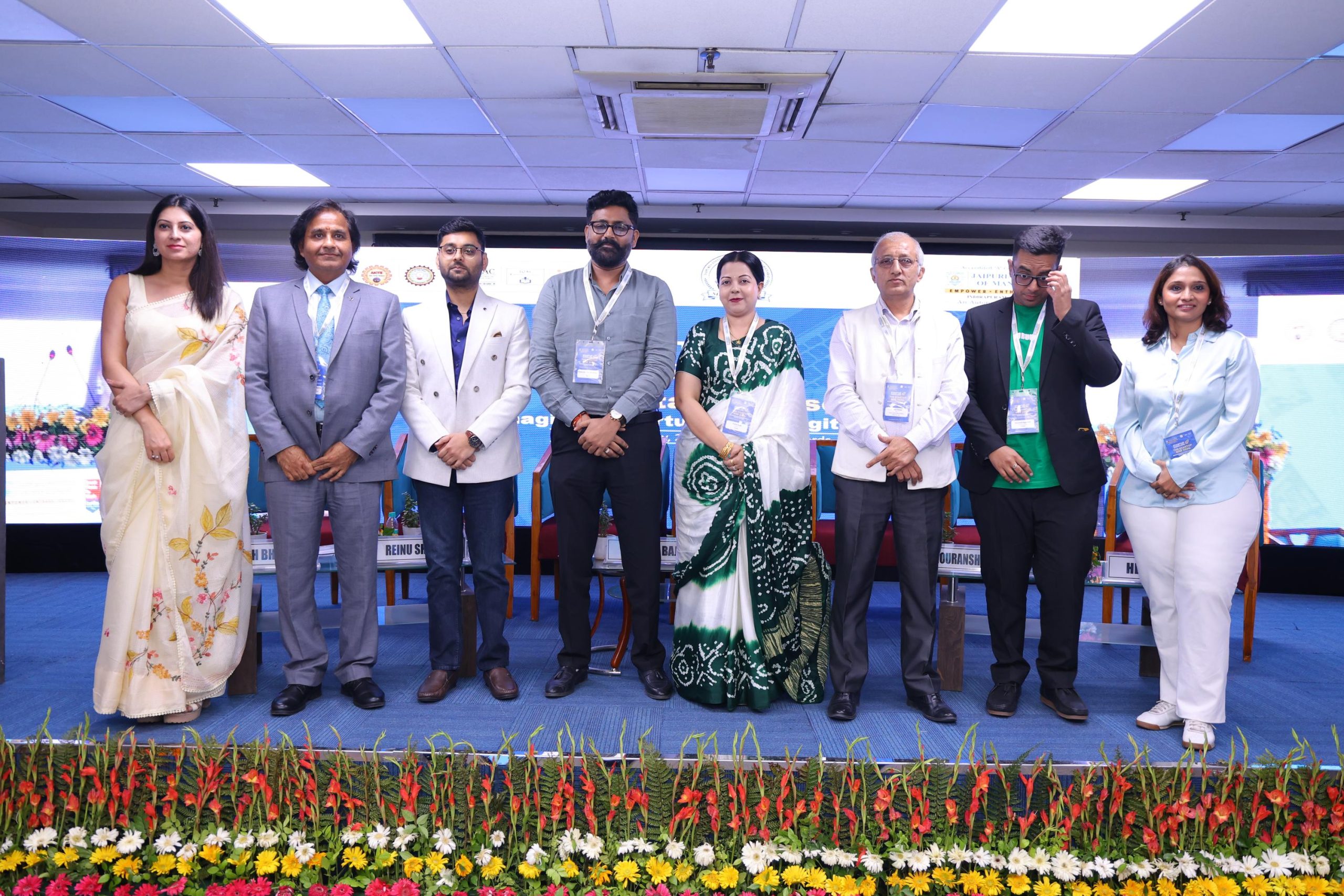 Startup Conclave 2026 Building Responsible Investment Ready Startups in the Digital Era 24 - Jaipuria Institute of Management, Indirapuram Ghaziabad Eight professionals in business attire pose on a conference stage, with a flower-filled foreground and banners in the background.