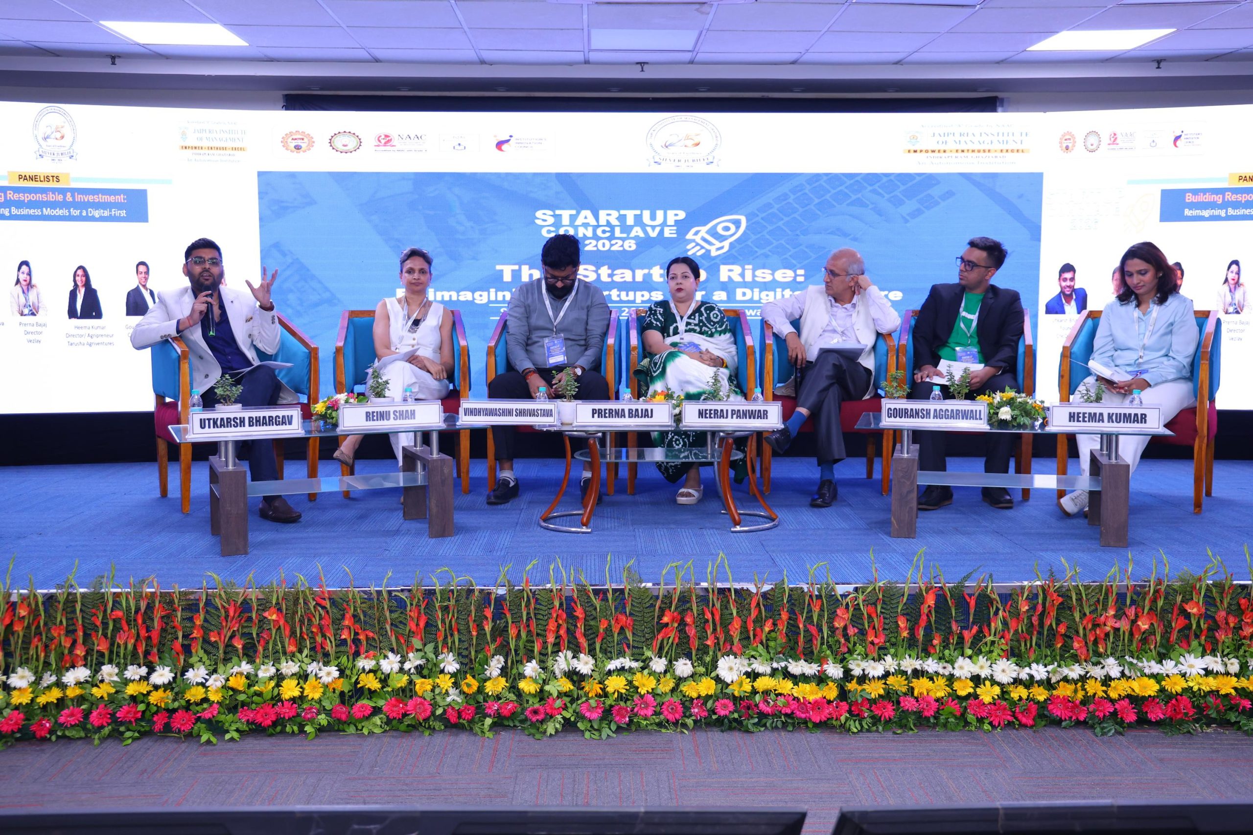 Startup Conclave 2026 Building Responsible Investment Ready Startups in the Digital Era 25 - Jaipuria Institute of Management, Indirapuram Ghaziabad Seven panelists seated in blue chairs on a stage, with a blue banner reading 'Startup Connect Live 2026' and a vibrant flower border in front.