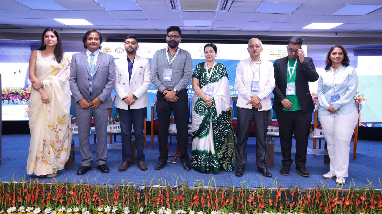 Nine professionals stand on a conference stage with lanyards, posing for a group photo in front of a blue backdrop and a flowered border at the front.