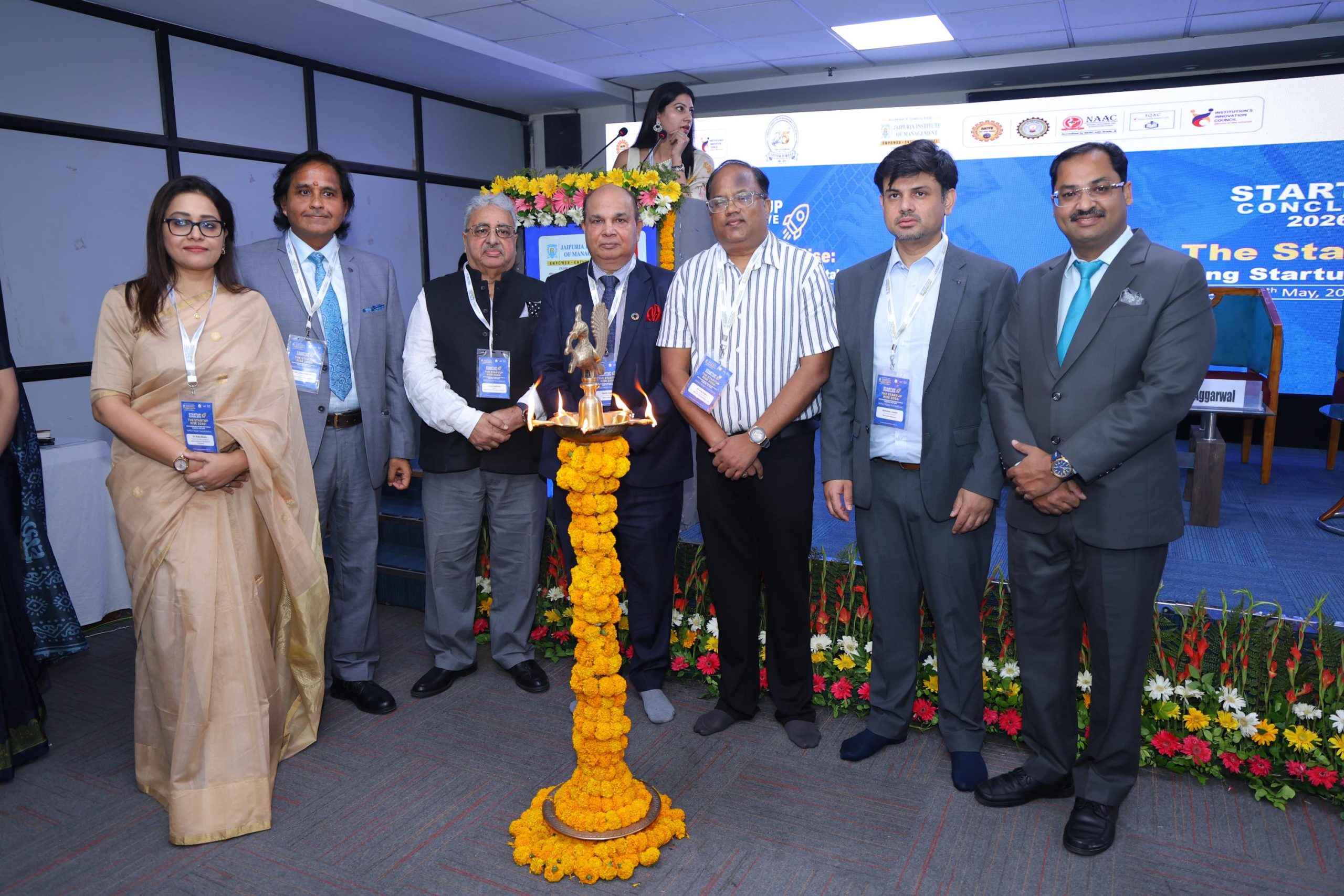Startup Conclave 2026 Guest of Honours 1 - Jaipuria Institute of Management, Indirapuram Ghaziabad Group of professionals in formal attire posing around a lit traditional lamp on a flower garland centerpiece during a conference ceremony.
