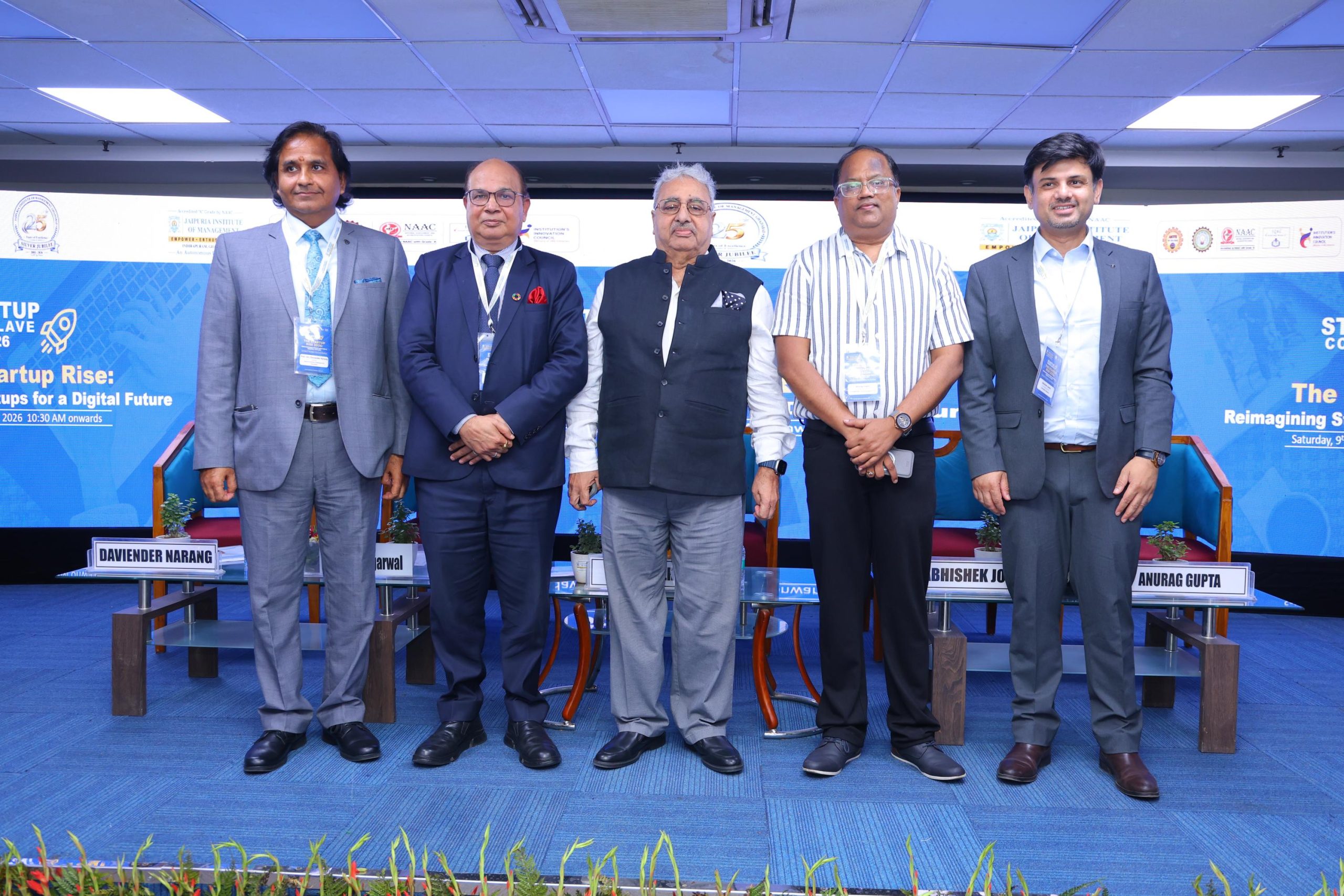 Startup Conclave 2026 Guest of Honours 17 - Jaipuria Institute of Management, Indirapuram Ghaziabad Five men in suits stand on a conference stage posing for a group photo, with blue banners and nameplates visible in front.