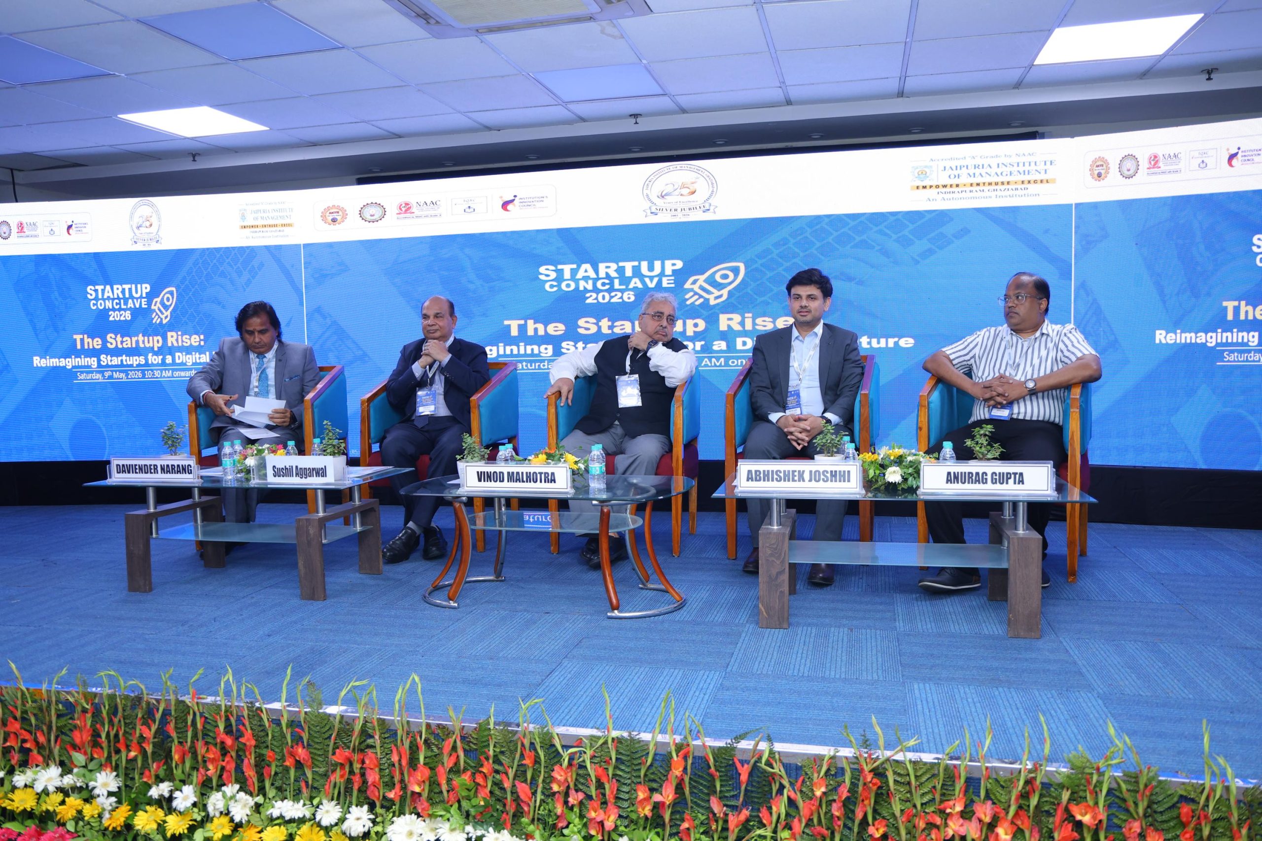 Startup Conclave 2026 Guest of Honours 7 - Jaipuria Institute of Management, Indirapuram Ghaziabad Panel of five men in suits seated on a blue conference stage with floral foreground and nameplates, during Startup Conclave 2026.