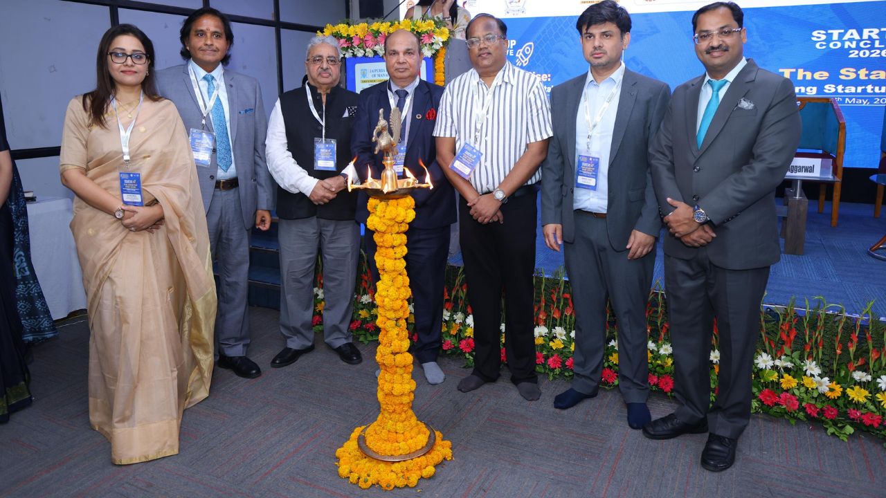 Group of professionals in formal attire around a flower-decorated lamp lighting ceremony at a conference stage, with name badges visible on their lanyards.