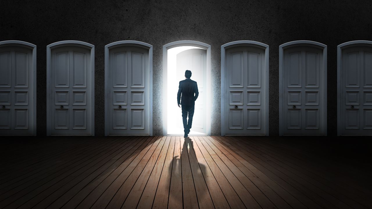Silhouette of a man in a suit walking through a bright open door in a row of closed doors, casting a long shadow.