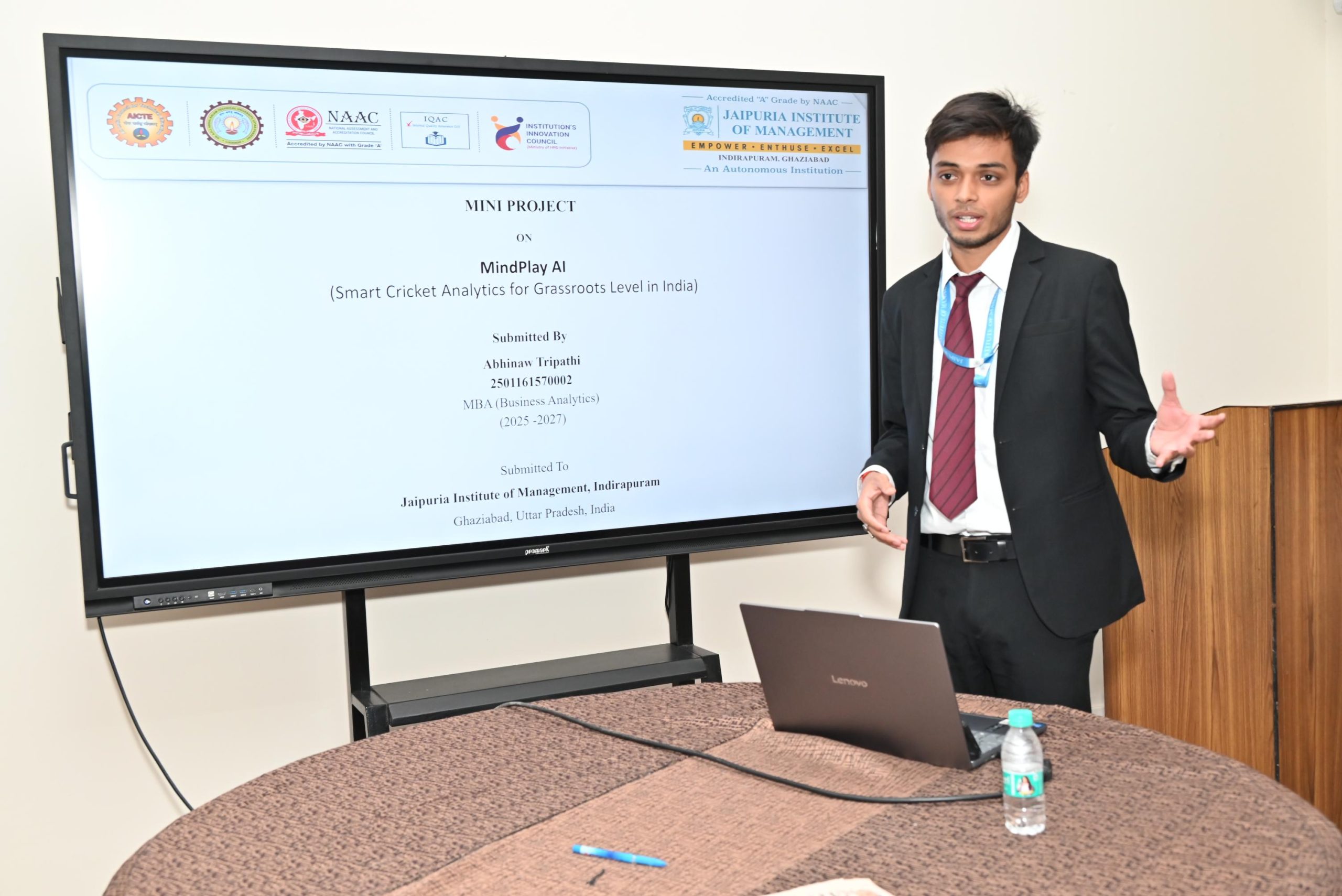 Workshop on Protecting IPR and IP Management for Startups 2026 1 - Jaipuria Institute of Management, Indirapuram Ghaziabad Presenter in a dark suit and red tie speaks beside a large screen displaying a slide titled 'MINI PROJECT' about MindPlay AI.