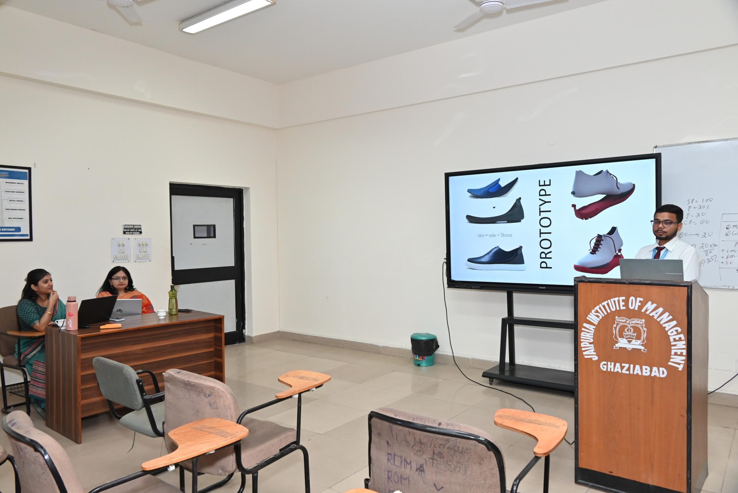 Workshop on Protecting IPR and IP Management for Startups 2026 2 - Jaipuria Institute of Management, Indirapuram Ghaziabad Classroom presentation with a speaker at a wooden podium and a large screen showing shoe designs; two women sit at a desk on the left.