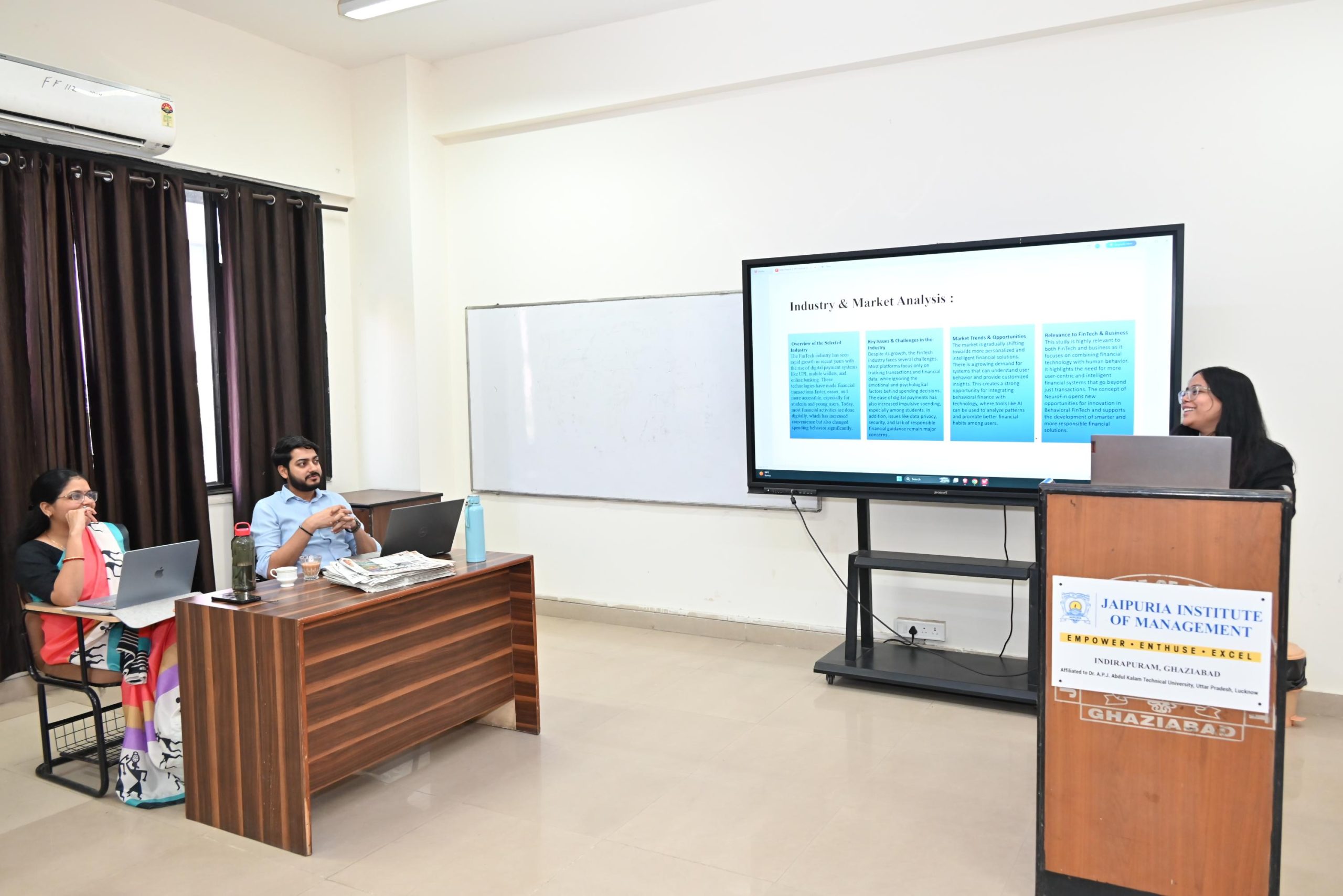 Workshop on Protecting IPR and IP Management for Startups 2026 5 - Jaipuria Institute of Management, Indirapuram Ghaziabad Group of students in a classroom watching a presenter at a podium with a large monitor displaying a slide on Industry & Market Analysis beside them at the front.