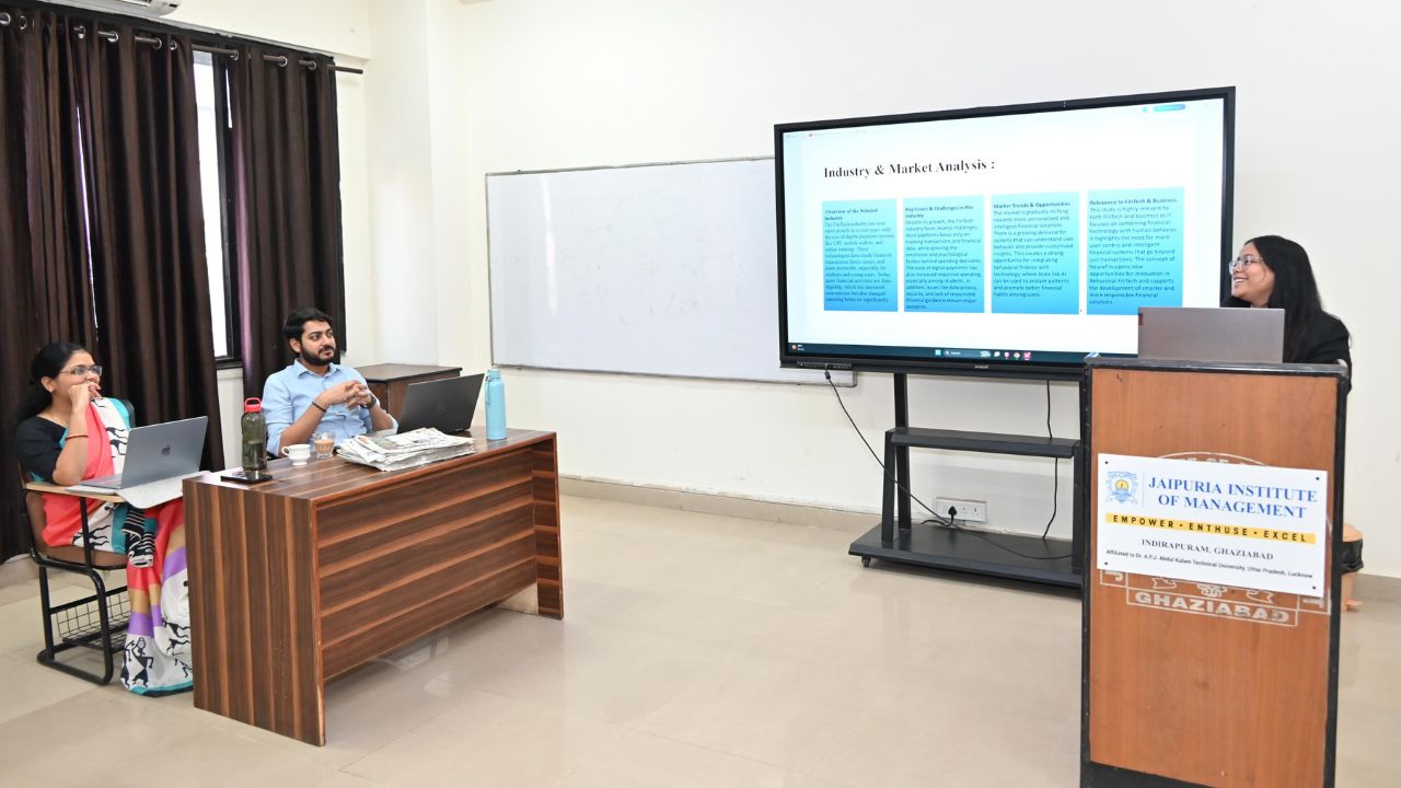 Female presenter at a podium showing a slide titled 'Industry & Market Analysis' to two seated students in a classroom.