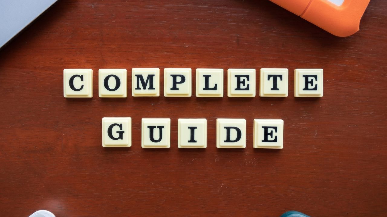 Scrabble tiles spell 'COMPLETE GUIDE' on a wooden desk, with parts of a laptop and an orange tape dispenser visible nearby.