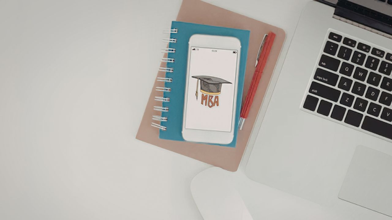 Desk setup with a laptop on the right, blue spiral notebook, a phone showing an MBA graduation cap illustration, and red pens nearby.