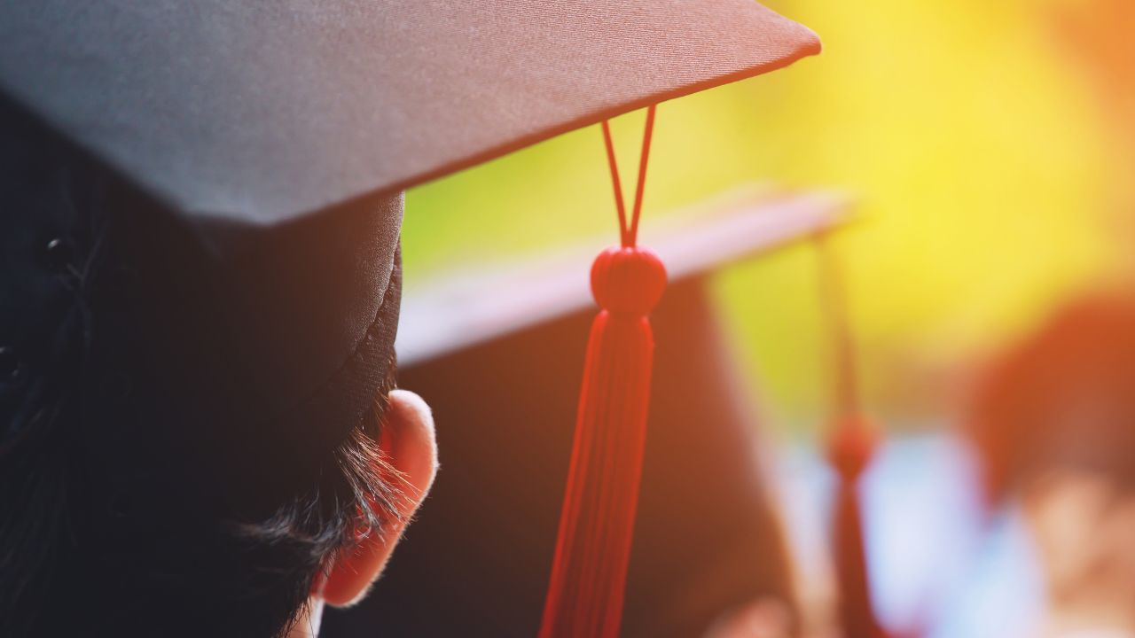 Close-up of a graduate's mortarboard with a red tassel, viewed from behind in warm sunlight.