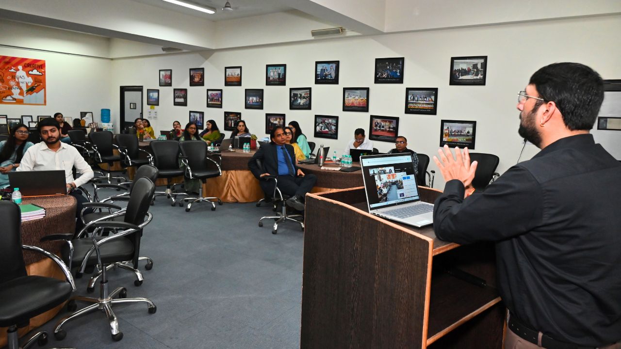 Professional meeting room: speaker at a podium presents to an audience around tables with laptops and water bottles on the desks, framed photos on the wall behind.