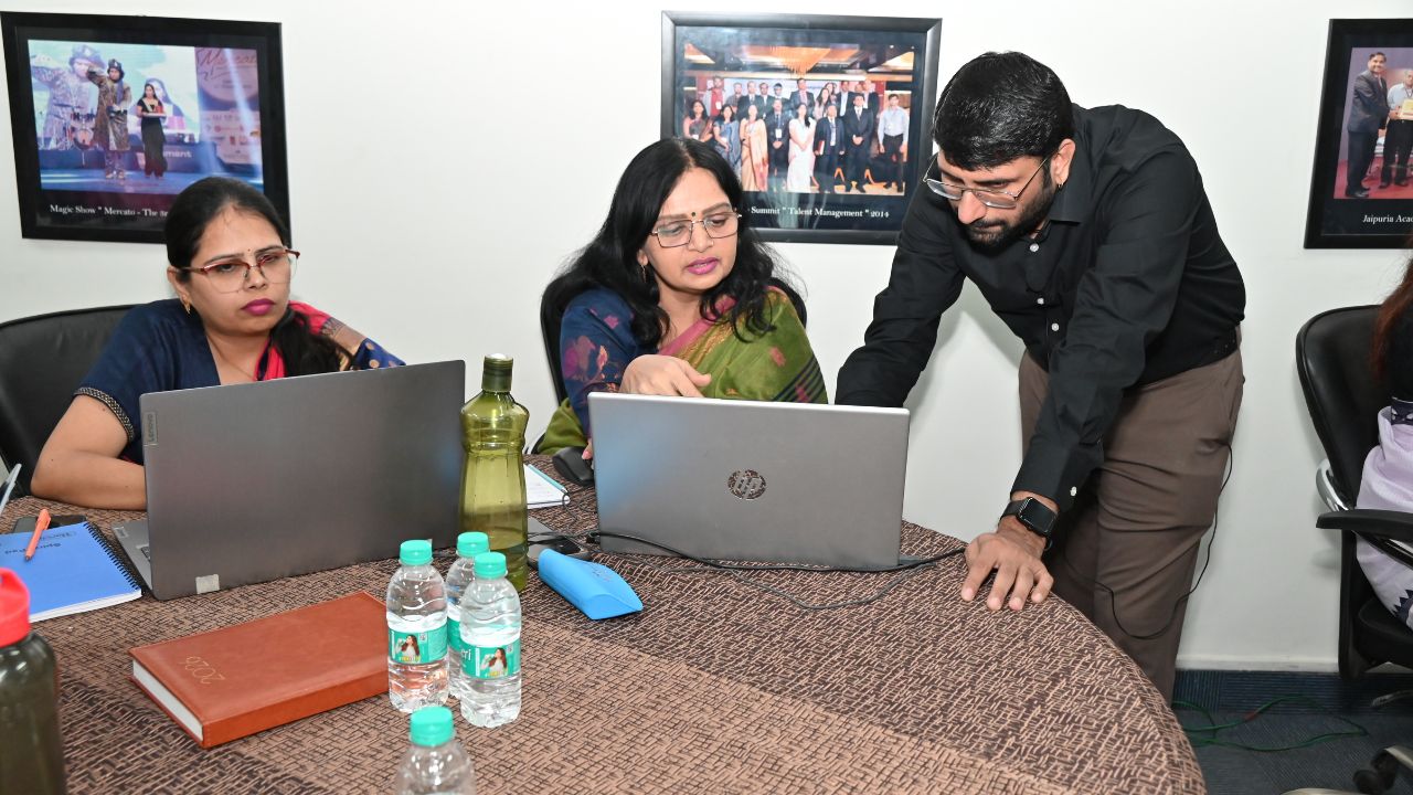 Report on Day 1 of the 5-Day Faculty Development Program 10 - Jaipuria Institute of Management, Indirapuram Ghaziabad Three colleagues work around a round table with laptops, one woman points at the screen while another watches intently as a man leans in to help.