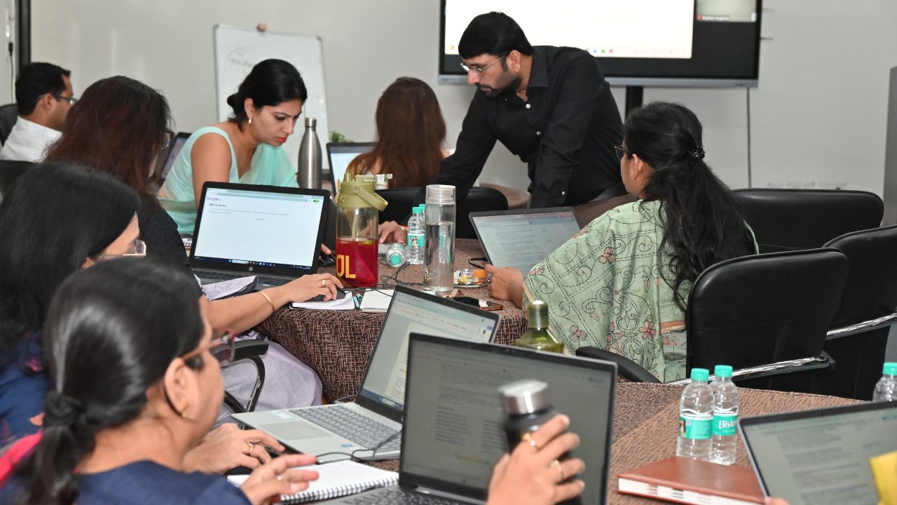 Report on Day 1 of the 5-Day Faculty Development Program 11 - Jaipuria Institute of Management, Indirapuram Ghaziabad Team of professionals around a conference table with laptops, water bottles, and a man leaning over to discuss something with a woman.