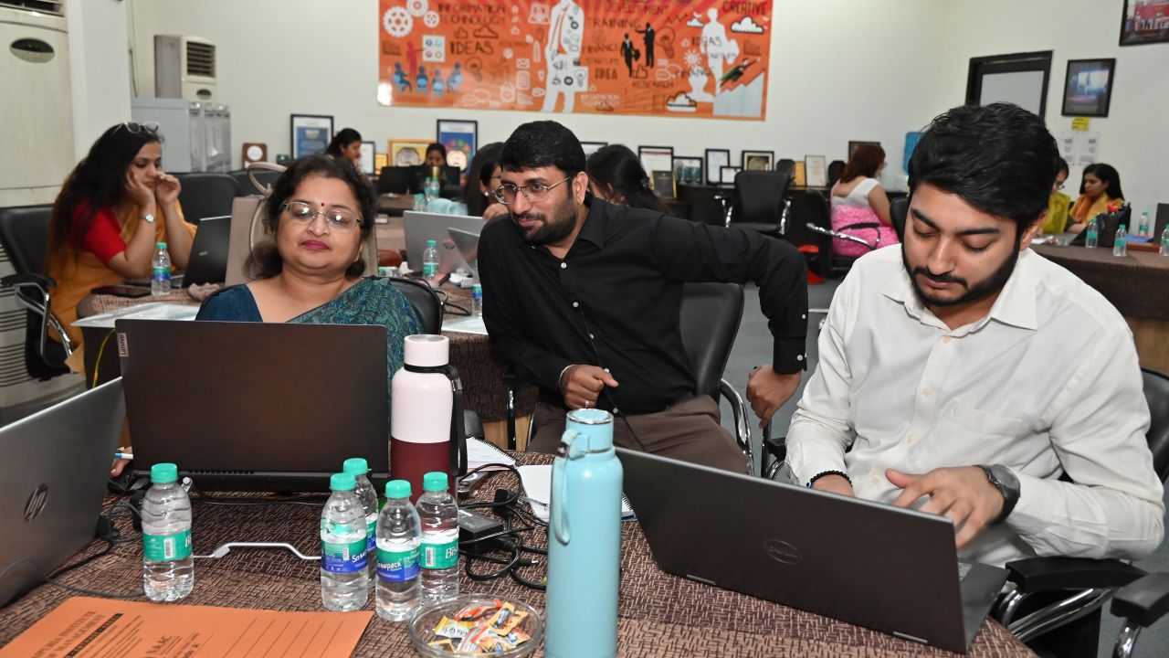 Report on Day 1 of the 5-Day Faculty Development Program 12 - Jaipuria Institute of Management, Indirapuram Ghaziabad Three colleagues work on laptops at a round table in a busy coworking space, with water bottles and a snacks dish in view.