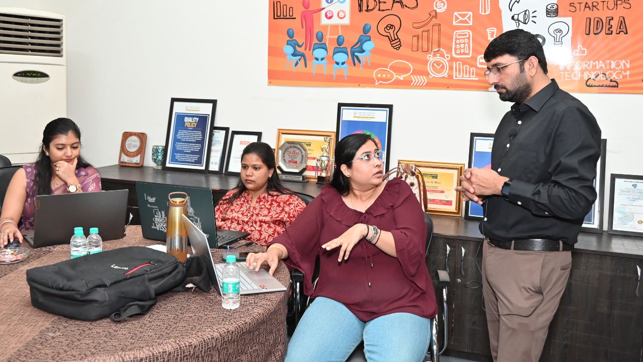 Report on Day 1 of the 5-Day Faculty Development Program 13 - Jaipuria Institute of Management, Indirapuram Ghaziabad Group of professionals in a meeting room; a man stands and speaks to a woman in a wheelchair while others work on laptops nearby.