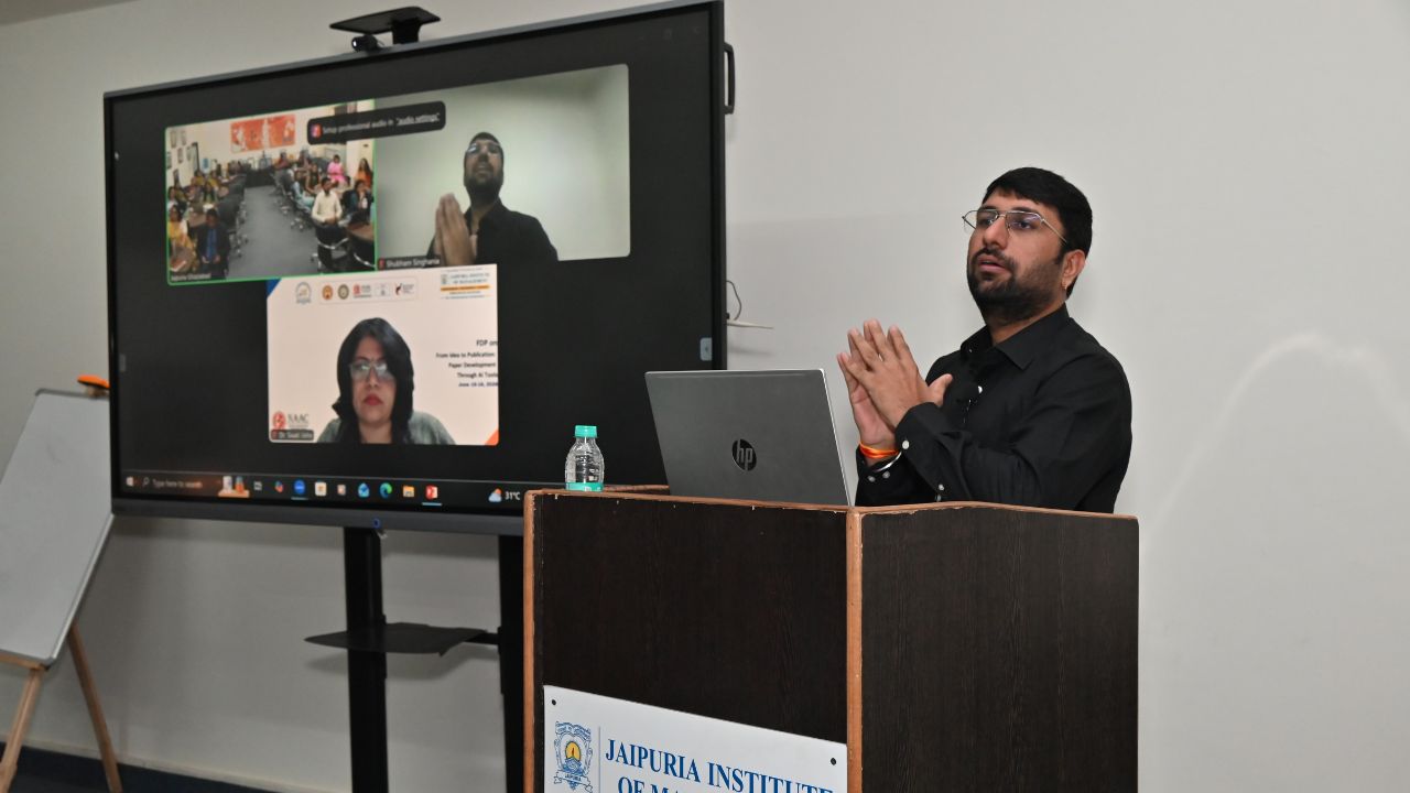 Report on Day 1 of the 5-Day Faculty Development Program 6 - Jaipuria Institute of Management, Indirapuram Ghaziabad Male presenter in a black shirt at a wooden podium with an open laptop; a large screen shows a hybrid meeting with remote participants.