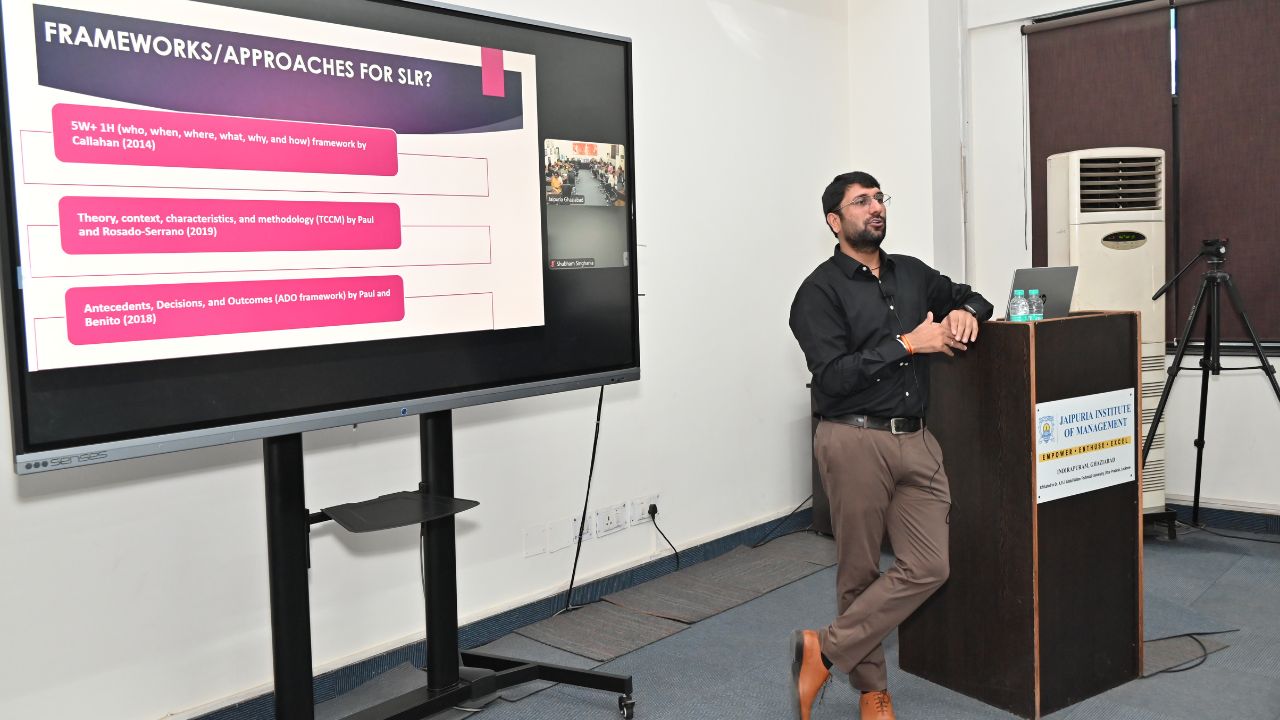 Report on Day 1 of the 5-Day Faculty Development Program 8 - Jaipuria Institute of Management, Indirapuram Ghaziabad Presenter at a podium beside a screen displaying a slide titled 'FRAMEWORKS/APPROACHES FOR SLR?' with pink bullet points.