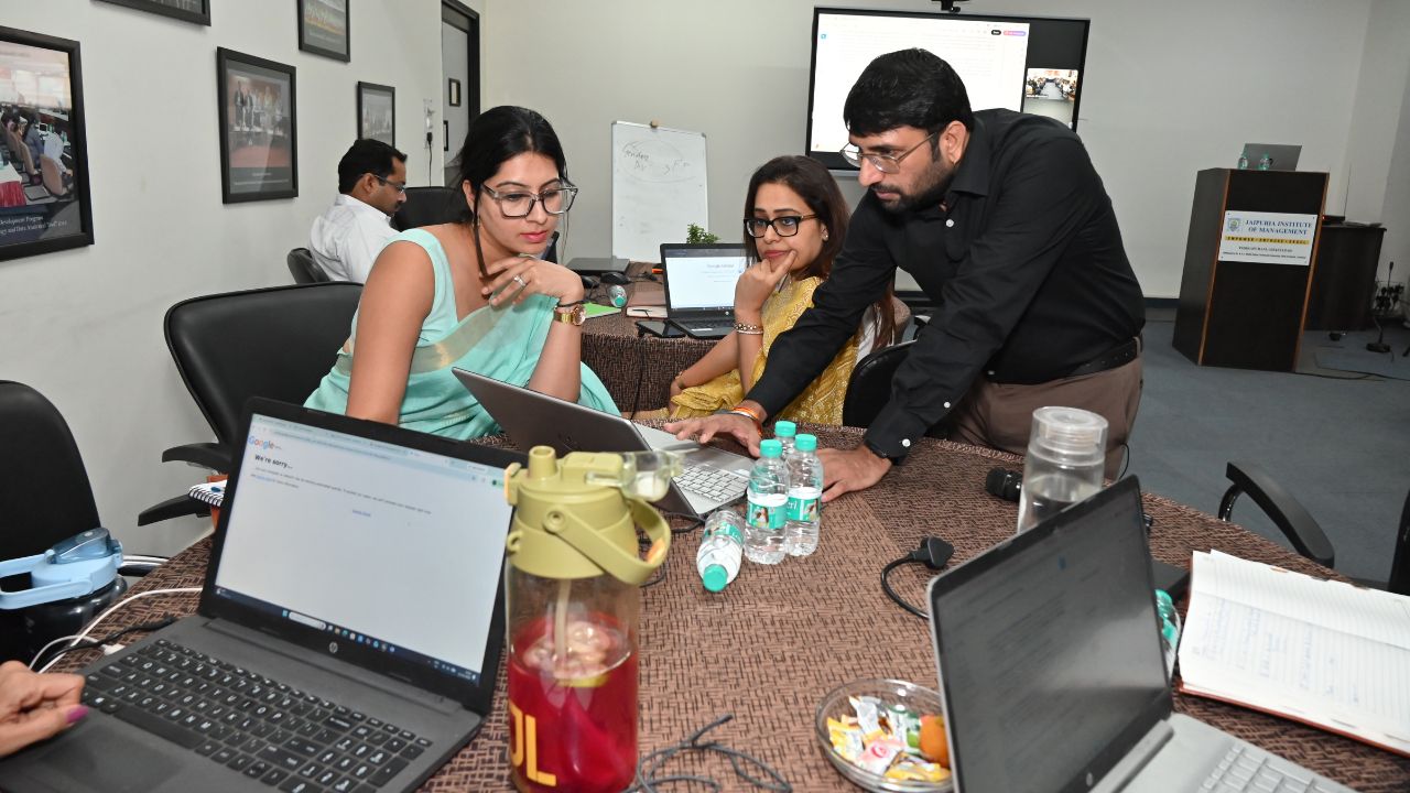 Report on Day 1 of the 5-Day Faculty Development Program 9 - Jaipuria Institute of Management, Indirapuram Ghaziabad Group of four colleagues around a conference table with laptops open; a man points at a screen as others look on.