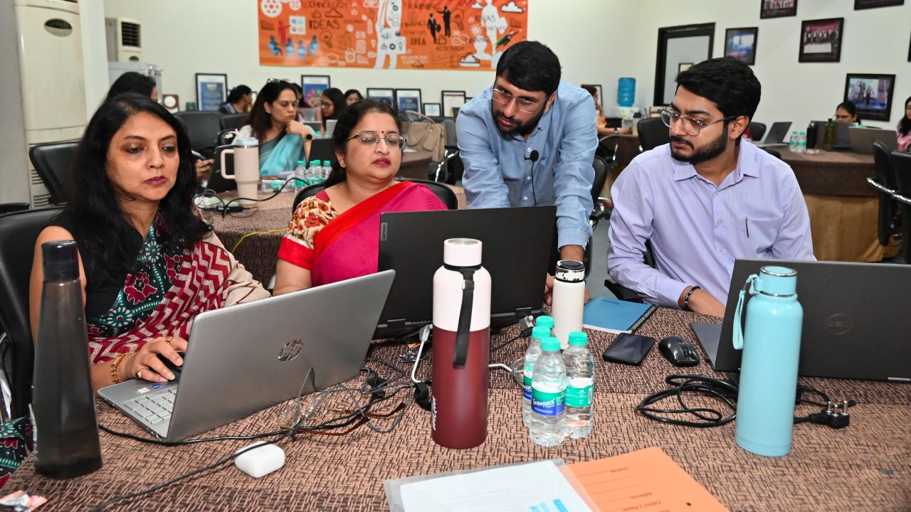 Report on Day 2 of the 5-Day Faculty Development Program 1 - Jaipuria Institute of Management, Indirapuram Ghaziabad Four colleagues collaborate around a table with laptops in a meeting room, discussing a laptop screen.