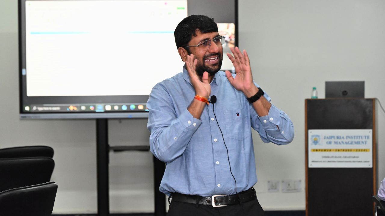 Report on Day 2 of the 5-Day Faculty Development Program 2 - Jaipuria Institute of Management, Indirapuram Ghaziabad Bearded man in a blue shirt speaking at a seminar, gesturing with his hands and wearing a lapel microphone in front of a projector screen.