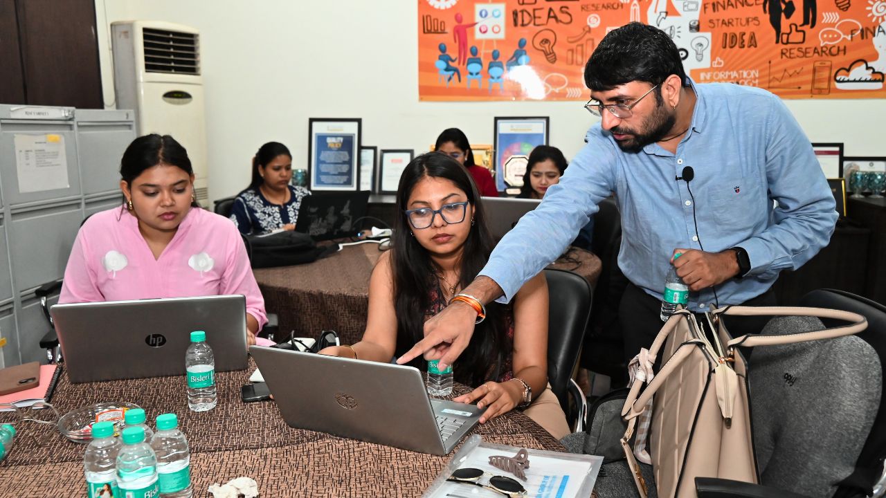 Report on Day 2 of the 5-Day Faculty Development Program 4 - Jaipuria Institute of Management, Indirapuram Ghaziabad Instructor in a blue shirt guides a student at a laptop during a workshop; others work on laptops around the table with water bottles nearby.