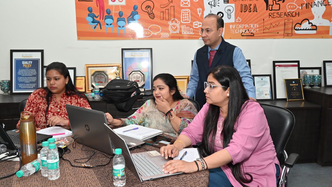 Report on Day 4 of the 5-Day Faculty Development Program 1 - Jaipuria Institute of Management, Indirapuram Ghaziabad Four professionals sit around a conference table with laptops and notebooks; a man stands behind them in an office setting, engaged in discussion.