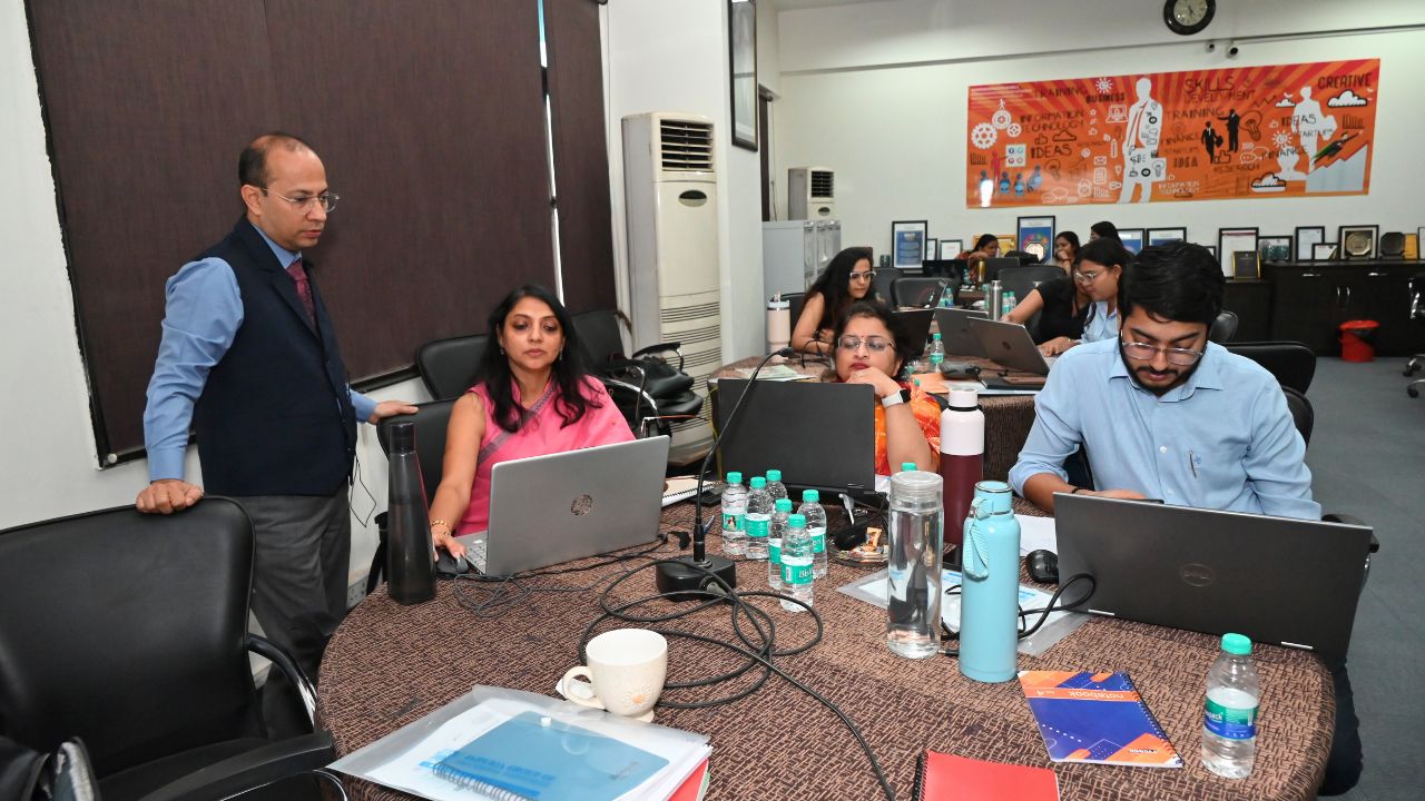 Report on Day 4 of the 5-Day Faculty Development Program 3 - Jaipuria Institute of Management, Indirapuram Ghaziabad Team of professionals in a training session around a round table, laptops open and water bottles on the table, facilitator standing nearby.