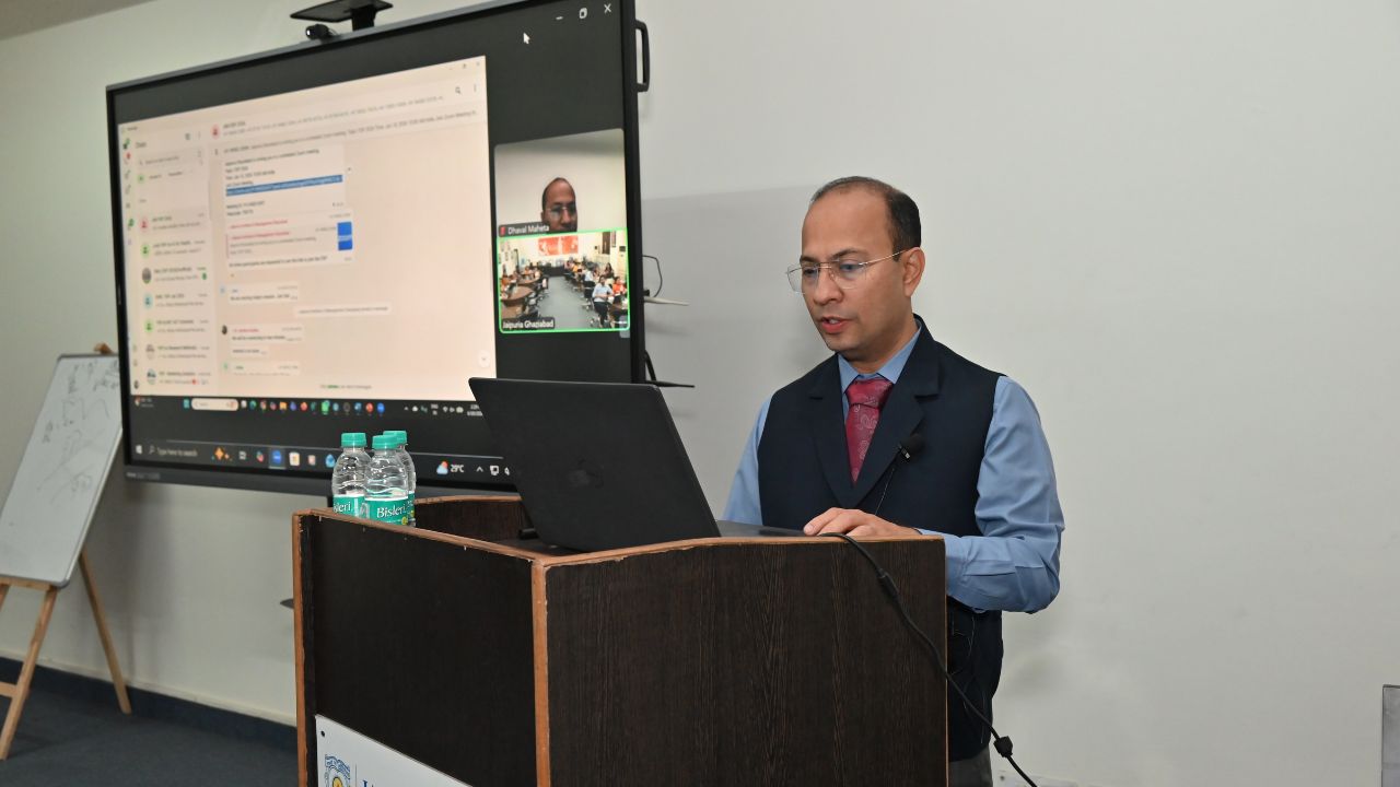 Report on Day 4 of the 5-Day Faculty Development Program 4 - Jaipuria Institute of Management, Indirapuram Ghaziabad Male presenter in a blue shirt and vest speaks at a lectern with a laptop; a large screen shows a video conference.