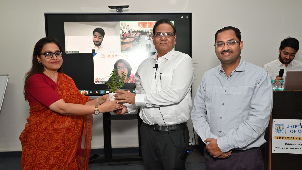 Report on Day 5 and Valedictory Session of the 5-Day Faculty Development Program 2 - Jaipuria Institute of Management, Indirapuram Ghaziabad Woman in a red sari hands a small potted plant to a man in a white shirt during a ceremony; a video call is visible on a screen in the background.