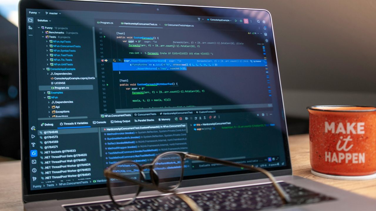 Open laptop showing a dark code editor with C# code, glasses resting on the keyboard, and a red mug that says "MAKE it HAPPEN" on a wooden desk.