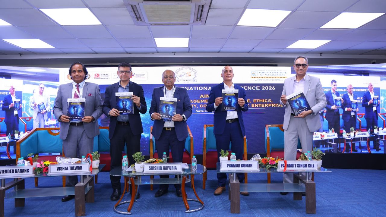 AIM-PEACE 2026_ International AI Conference at Jaipuria MBA 1 - Jaipuria Institute of Management, Indirapuram Ghaziabad Five men in suits stand on a conference stage, each holding a blue booklet; nameplates with attendee names sit on the table in front.