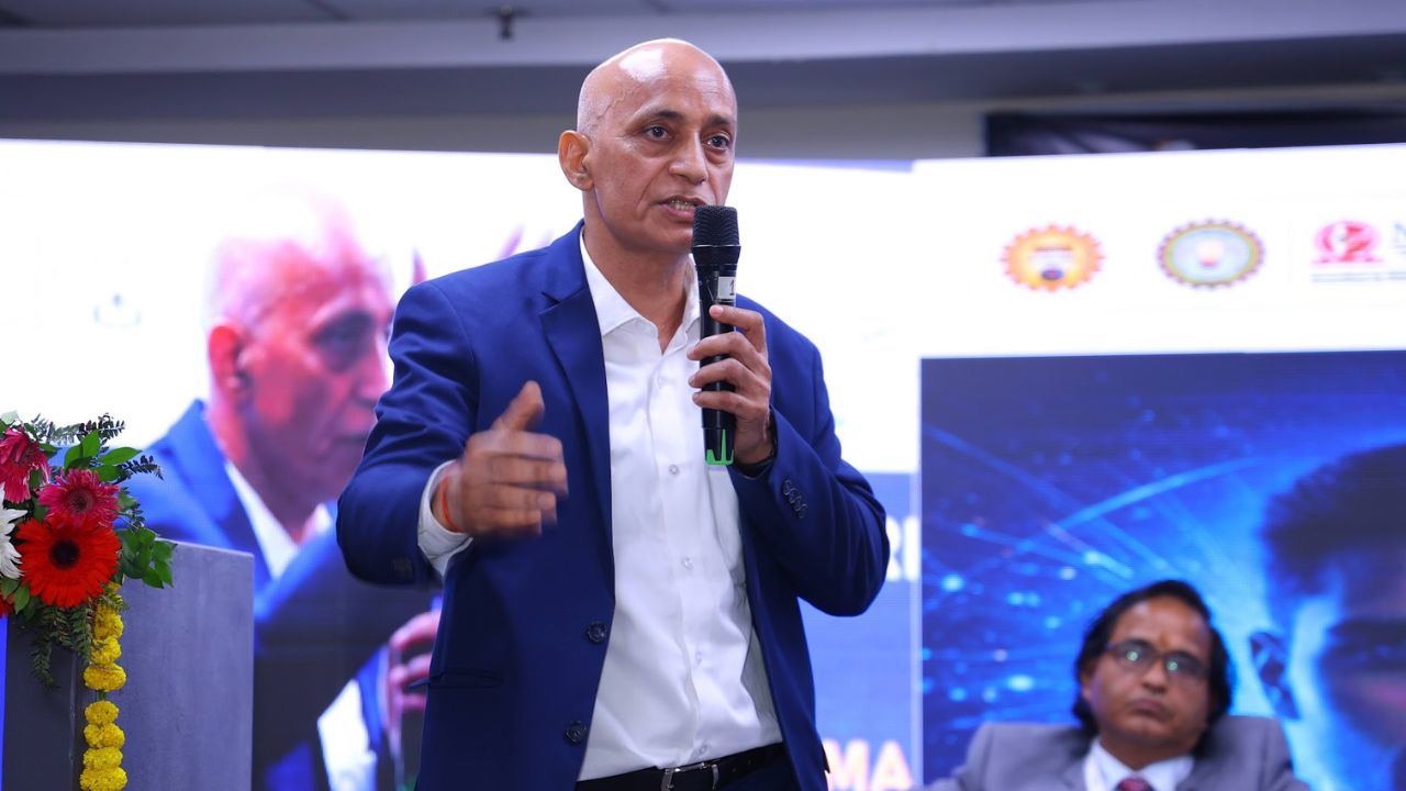 AIM-PEACE 2026_ International AI Conference at Jaipuria MBA 7 - Jaipuria Institute of Management, Indirapuram Ghaziabad Man in a blue blazer speaks into a handheld microphone on stage, gesturing with his other hand; a large screen with blue graphics is behind him.
