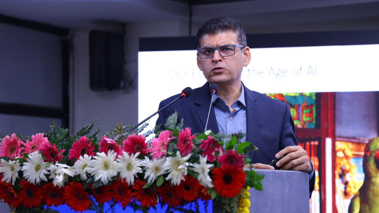 AIM-PEACE 2026_ International AI Conference at Jaipuria MBA 8 - Jaipuria Institute of Management, Indirapuram Ghaziabad Man in a blue suit and glasses speaks at a podium with dual microphones, in front of a lush floral arrangement of pink, red, and white flowers.