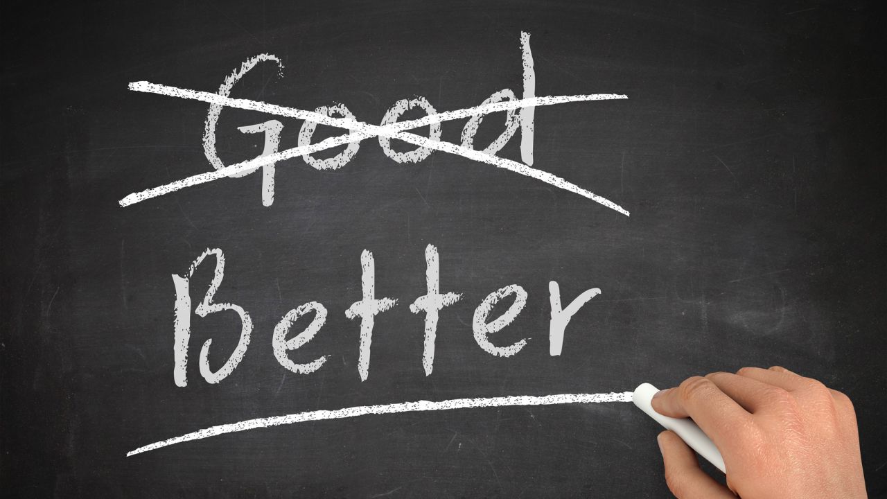 Hand holding chalk on a chalkboard, with the word 'Good' crossed out and 'Better' written underneath with an underline, signaling improvement.