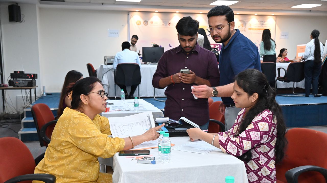 Day 1 of Reporting Day 1 - Jaipuria Institute of Management, Indirapuram Ghaziabad Two women seated at a table during an event exchange papers and discuss documents, with water bottles and pens on the table.