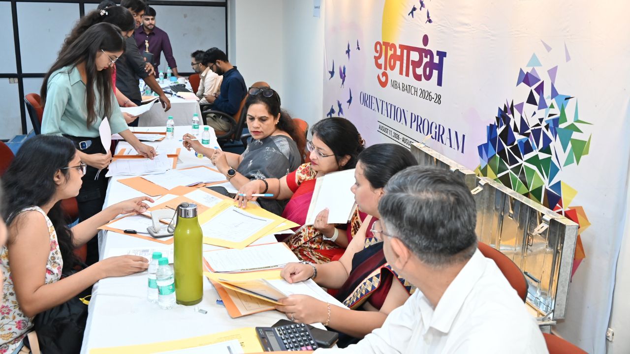 Day 1 of Reporting Day 2 - Jaipuria Institute of Management, Indirapuram Ghaziabad People sit around a long registration table with papers, pens, and water bottles during an orientation program banner behind them