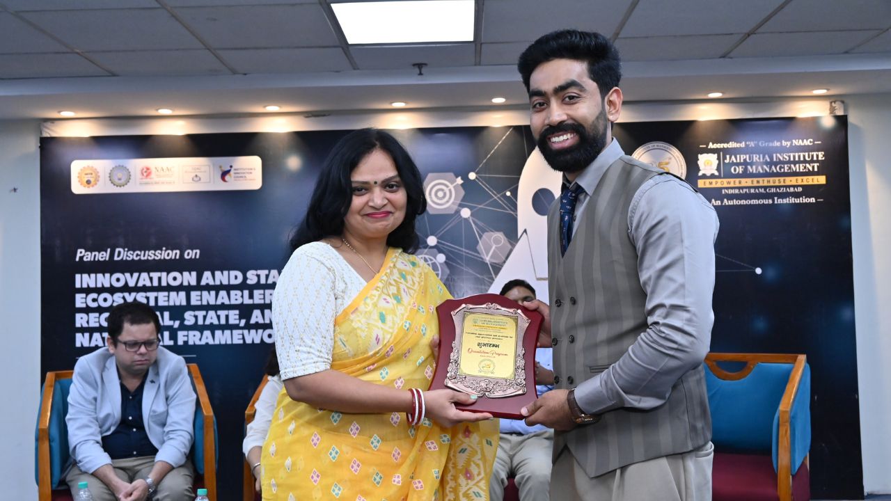 IIC Panel Discussion on Innovation & Startup Ecosystem (10) - Jaipuria Institute of Management, Indirapuram Ghaziabad Man in a suit hands a plaque to a woman in a yellow saree at a panel discussion event, both smiling, backdrop shows conference banners.