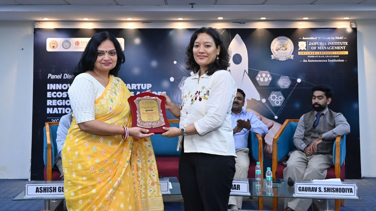 IIC Panel Discussion on Innovation & Startup Ecosystem (11) - Jaipuria Institute of Management, Indirapuram Ghaziabad Two women exchange a framed award on stage during a panel discussion, with a dark banner and seated panelists in the background.