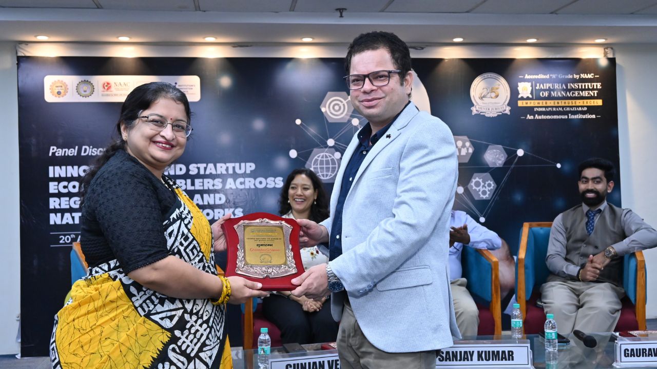 IIC Panel Discussion on Innovation & Startup Ecosystem (12) - Jaipuria Institute of Management, Indirapuram Ghaziabad Woman receives a plaque from a man on stage during an award ceremony at a startup panel discussion event.
