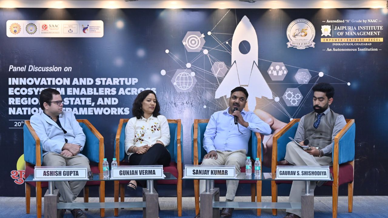 IIC Panel Discussion on Innovation & Startup Ecosystem (8) - Jaipuria Institute of Management, Indirapuram Ghaziabad Panel discussion with four speakers seated in colorful chairs on a stage, nameplates read Ashish Gupta, Gunjan Verma, Sanjay Kumar, and Gaurav S. Shishodiya, backdrop about innovation and startup ecosystems.