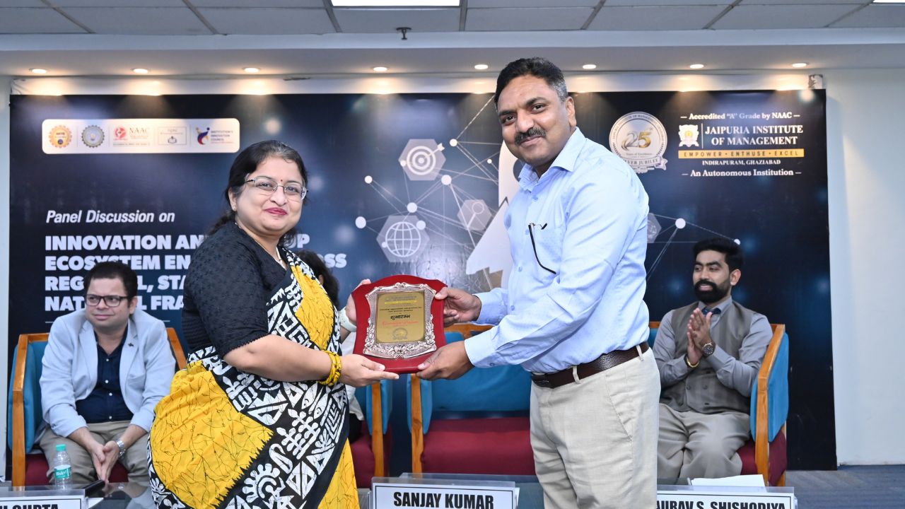 IIC Panel Discussion on Innovation & Startup Ecosystem (9) - Jaipuria Institute of Management, Indirapuram Ghaziabad Man in blue shirt hands plaque to woman in yellow and black sari on stage during award ceremony; panel backdrop and seated guests in background.